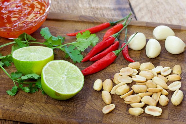 Ingredients on a wooden board for making a spicy sauce for mangos including peanuts, Thai red chili peppers, garlic cloves and cilantro sprigs with a glass bowl of ready-made sauce on the side.