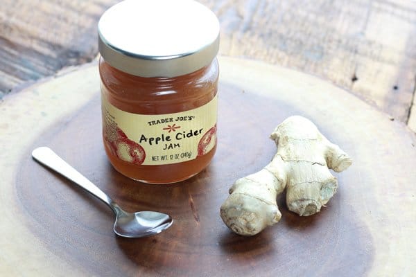A glass jar of Trader Joe's apple cider jam on top of a wooden board with a spoon and a ginger bulb on either side.