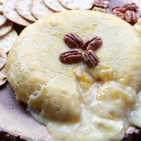 A golden-brown baked brie oozing with cheese and topped with pecans right out of the oven placed on top of a wooden board with crackers and pecans placed behind.