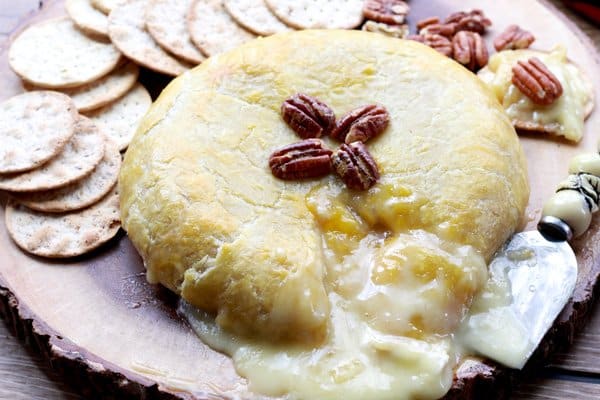 A round brie baked in puff pastry with melty cheese oozing out on top of a wooden board with crackers along side.