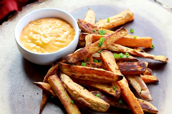 Baked sweet potato fries piled on a wooden board with a small white bowl of sriracha aioli dipping sauce on the side.