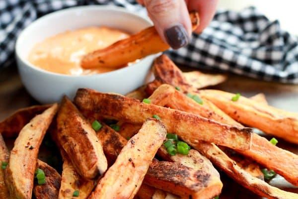 A stack of sweet potato fries on a plate with a woman's hand dipping a fry into a small white bowl of aioli.