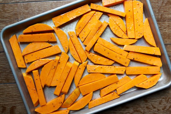 A baking sheet lined with thickly sliced butternut squash sprinkled with salt and pepper on top of a wooden board ready for the oven.