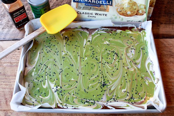 matcha holiday bark on a tray topped with sesame seeds with bags of chocolate chips and a rubber yellow spatula on the side