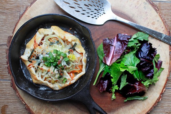 A butternut squash galette baked inside a mini cast iron skillet placed on top of a wooden board with a colorful wild greens salad on the side.