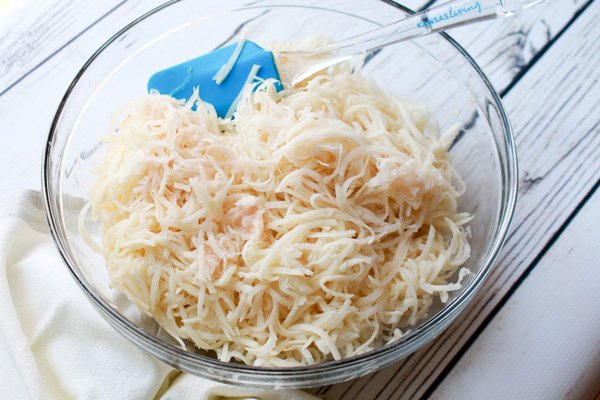 Shredded potatoes in a glass mixing bowl on top of a white wooden board.