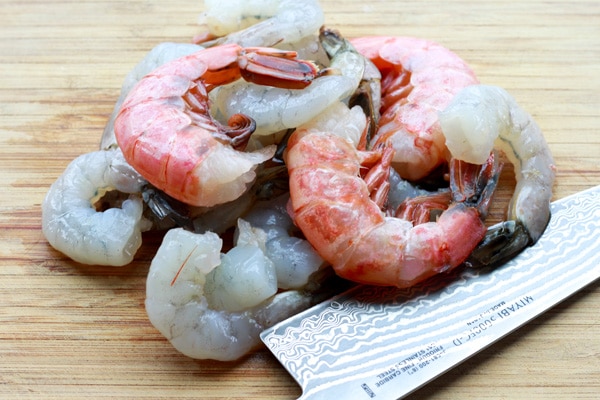 A pile of raw shrimp placed on a wooden cutting board with a chef's knife on the side.