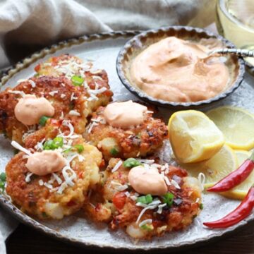 Golden-brown and crispy coconut shrimp cakes on a round white serving plate with a spicy aioli sauce, lemon wedges, and hot red peppers on this side and a glass of white wine in the background.