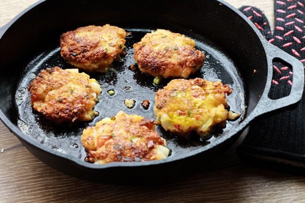 Golden-brown coconut shrimp cakes frying up in a cast iron skillet placed on top of a wooden board.