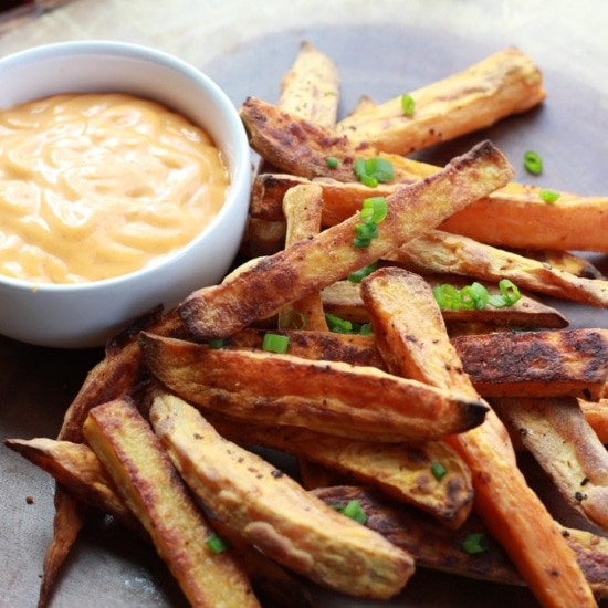 Baked sweet potato fries piled on a wooden board with a small white bowl of sriracha aioli dipping sauce on the side.