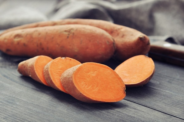 
Sliced sweet potatoes on a wooden board with two whole potatoes in the background.