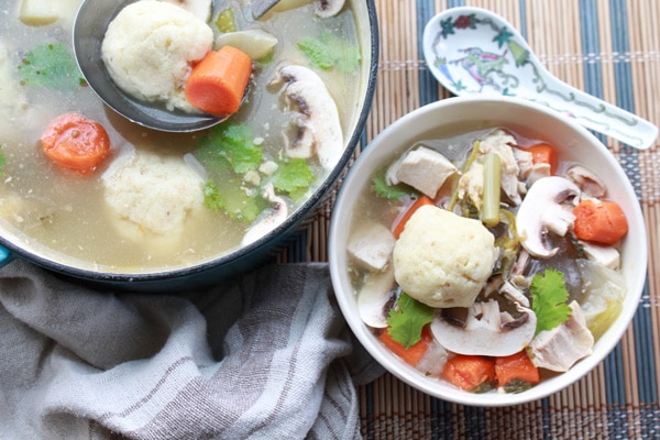 A white bowl of matzo ball soup with veggies on top and a soup spoon on the side with a pot of soup in the background placed on top of a bamboo placemat.