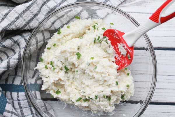Cauliflower mash with chives in a large glass mixing bowl and red spatula inserted placed on top of a white wooden board with a kitchen towel on the side.