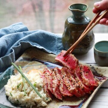 Slices of sesame seared tuna on a square plate with a side of cauliflower mash topped with green onions and a hand picking up a slice of tuna with chopsticks placed on a white board with a sake set and blue napkin in the background.