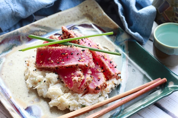 A square plate with slices of sesame seared tuna placed on top of cauliflower mash with a pair of chopsticks on the side.