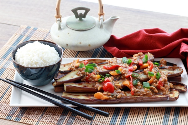 A long white narrow plate with charred Chinese eggplant topped with a garlic pepper sauce with a bowl of white rice and black chopsticks along side placed on top of a bamboo placemat with a red napkin and small teapot on the side.