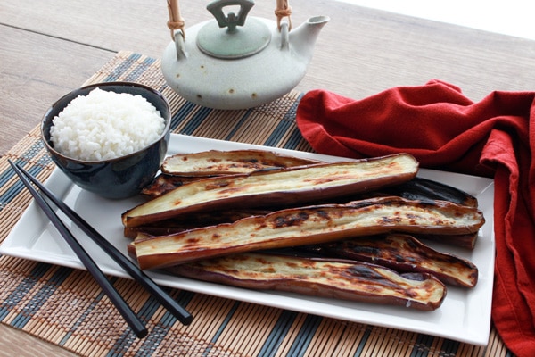 A long white narrow plate with charred Chinese eggplant with bowl of white rice and black chopsticks along side placed on top of a bamboo placemat with a red napkin and small teapot on the side.