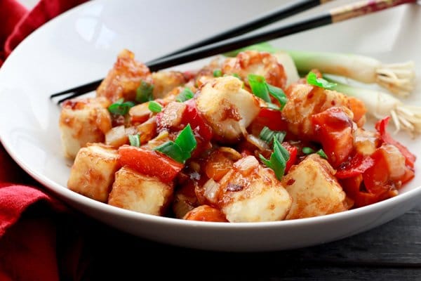 Crispy tofu cubes in a sweet and sour sauce in a white bowl with chopsticks and a red napkin on the side.