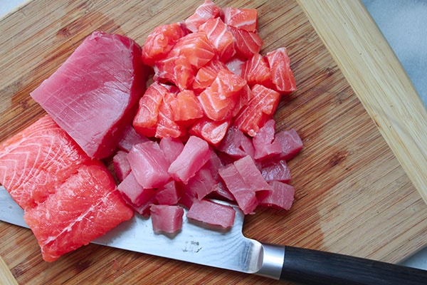 cubes of sushi-grade salmon and tuna on a wooden cutting board with a chef's knife on the side.