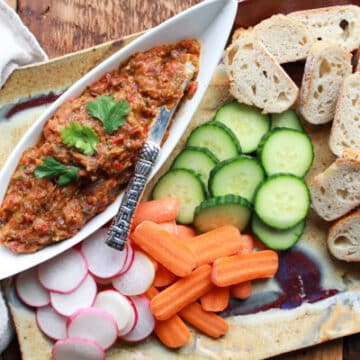 Roasted Chinese eggplant dip in a white boat-shaped bowl with fresh veggies and sliced bread on the side.