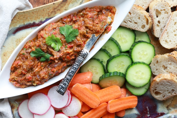 Roasted eggplant dip prepared in a white narrow bowl with fresh veggies and slices of bread on the side.