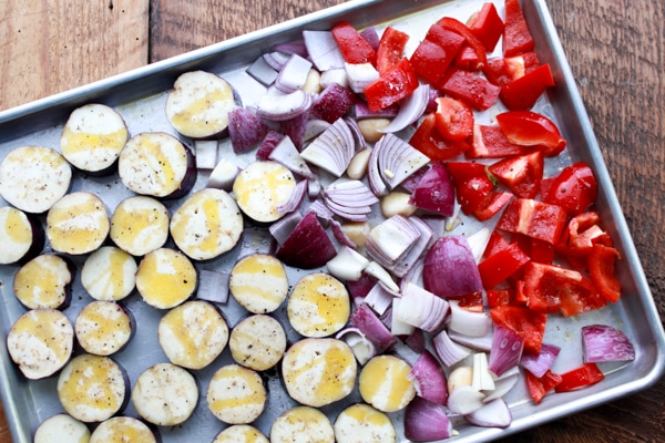 sliced eggplant, red onions, and red bell peppers on a baking sheet ready for the oven.