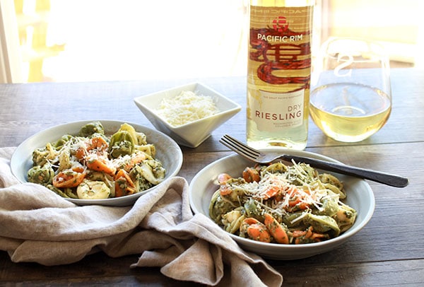 Tri-color tortellini in two white bowls tossed in a Thai pesto sauce with a fork on the side of the bowl and a glass and bottle of Reisling wine in the background.