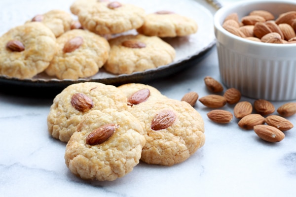 Chinese almond cookies piled on a round white plate with bowl of almonds on the side and extra cookies in front on top of a marble surface.