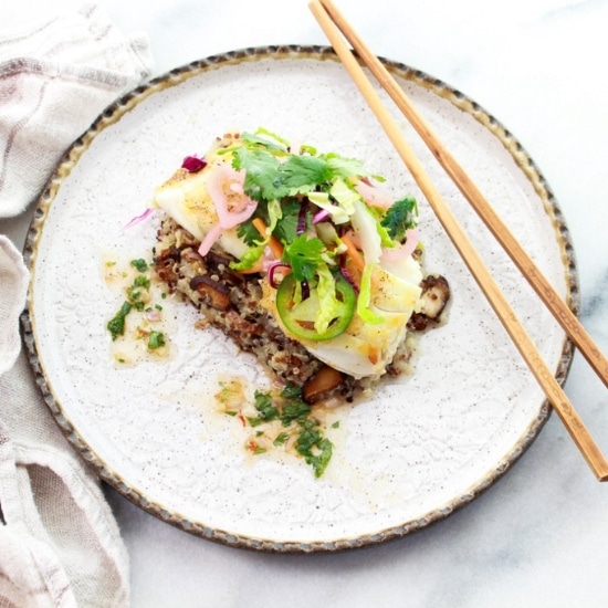 Pan seared halibut on top of a bed of quinoa and topped with a spicy nuoc cham slaw on top of a round white plate with chopsticks on the side.