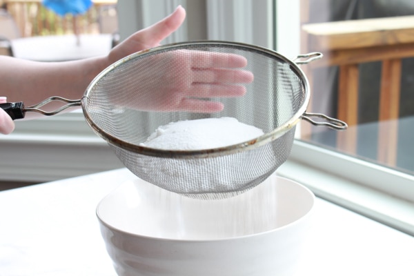 A woman's hand sifting flour in a sifter on top of a white bowl.