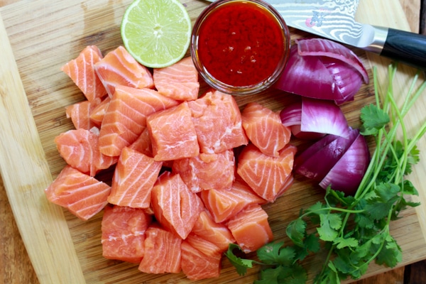 cubed raw salmon on a cutting board with lime, sauce, red onions, and cilantro sprigs