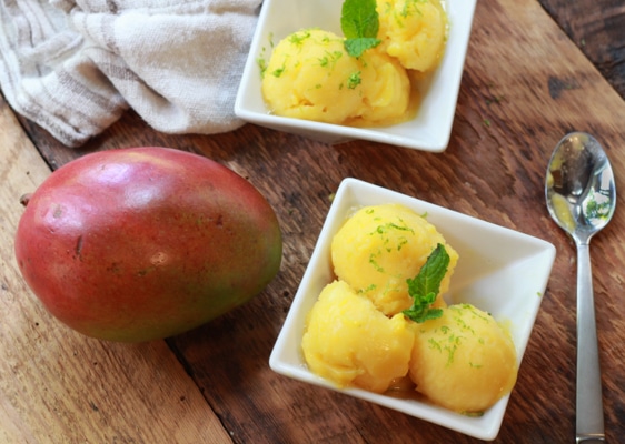 mango coconut sorbet in two white bowls with a spoon and whole mango on the side on top of a wooden board