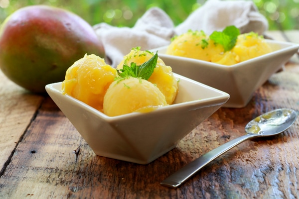 mango coconut sorbet in two white bowls with a spoon and whole mango on the side on top of a wooden board