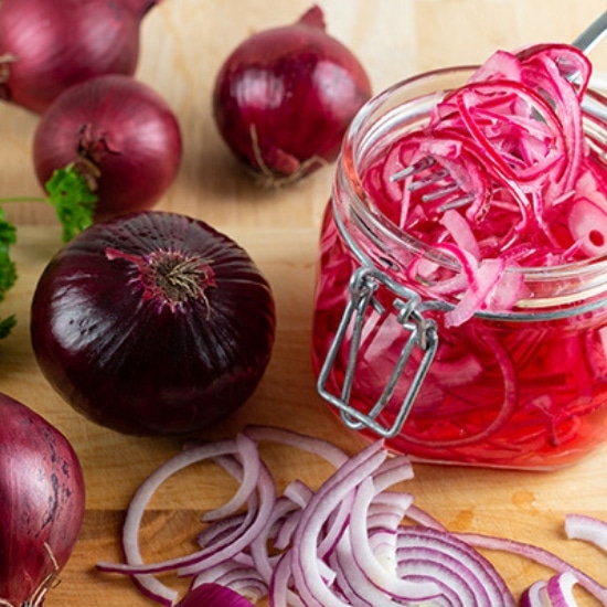 homemade sliced pickled red onions in a glass mason jar with raw onions on a wooden board