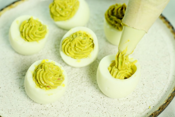 piping deviled egg mixture into an egg white on a white plate with other deviled eggs
