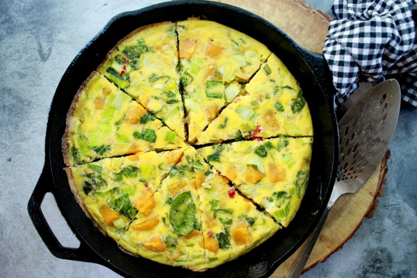 a roasted butternut squash and baby bok choy frittata in a cast iron pan on a round wooden board with a silver serving piece and checkered napkin