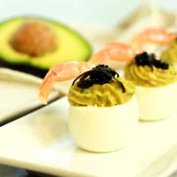 avocado deviled eggs topped with fresh shrimp and caviar stacked on a white plate with a halved avocado in the background.