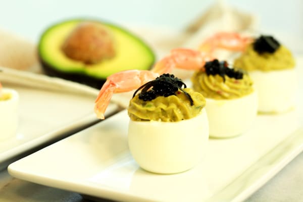 Avocado deviled eggs topped with fresh shrimp and black caviar stacked on a white plate with a halved avocado in the background.