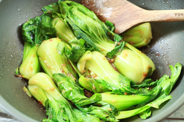 baby bok choy being stir-fried in a wok with sauce and a wooden spatula