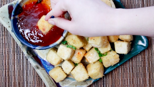 crispy tofu cubes on a plate with a woman's hand dipping one piece into a chili sauce