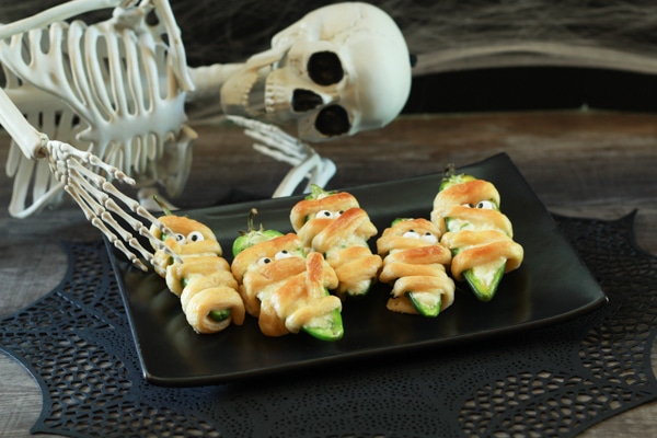 Halloween Jalapeño Poppers decorated like Mummies on a black plate with a skeleton laying beside it.