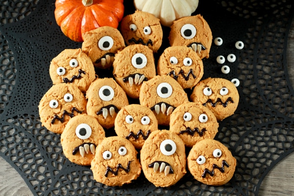 Cutely decorated pumpkin monster cookies on top of a black spiderweb place mat with baby pumpkins in the background.