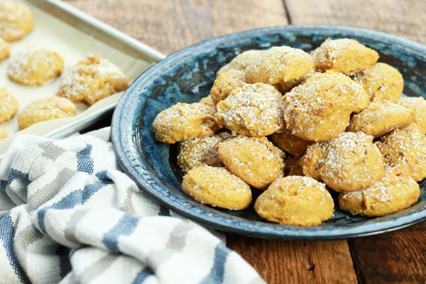 pumpkin spice cookies sprinkled with powdered sugar piled on a blue plate with extra cookies on a baking tray and a striped napkin on the side