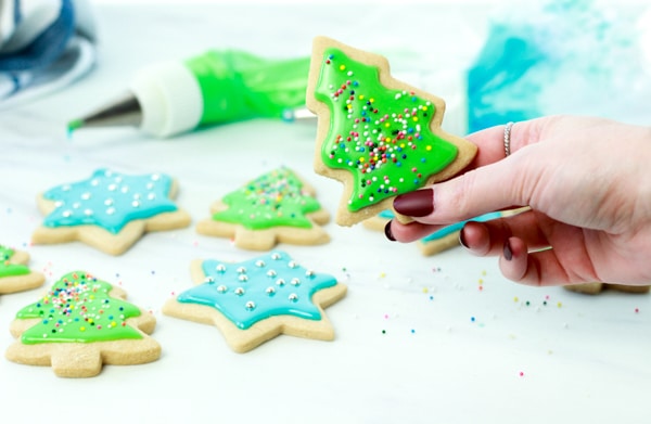woman holding up a sugar cookie with green glaze shaped like a Christmas tree with other holiday cookies underneath