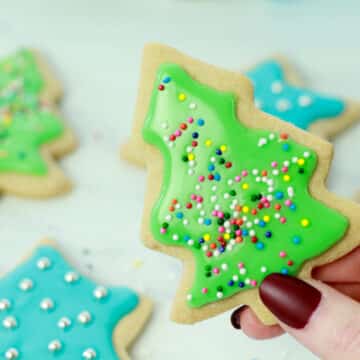 A woman's hand holding up a decorated green Christmas tree sugar cookie with other cookies in the background.