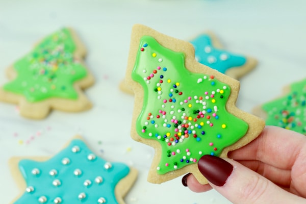 A woman's hand holding up a decorated green Christmas tree sugar cookie with other cookies in the background.