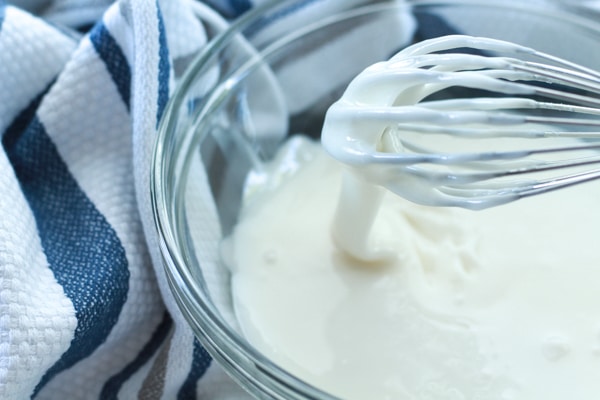 white frosting in a glass bowl with a whisk and a blue and white kitchen towel on the side