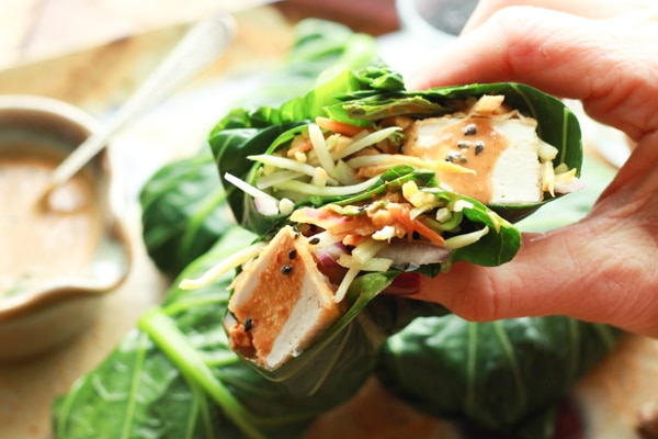 a hand holding a collard green wrap stuffed with tofu and broccoli slaw and topped with peanut sauce