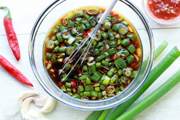 an Asian marinade in a glass bowl with a whisk with scallions, garlic cloves, and red peppers on the side