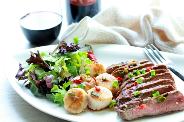 grilled flank steak slices and seared scallops on a white plate with a green salad on the side with a bottle and glass of red wine and napkin on the side